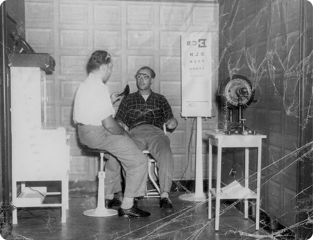 An optician examining a seated customer beside an eye chart and refracting equipment.