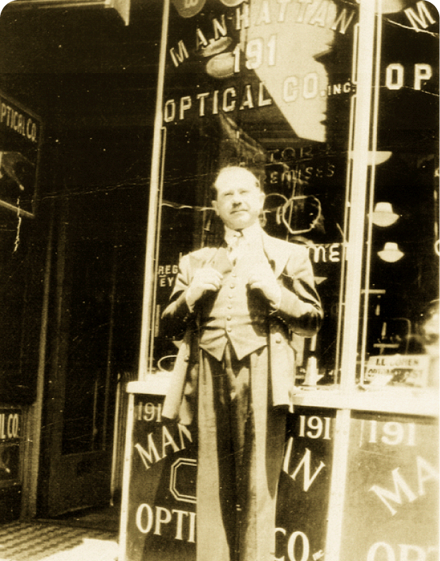 A moustached man in a three-piece suit standing outside the Manhattan Optical Co. storefront at number 191.