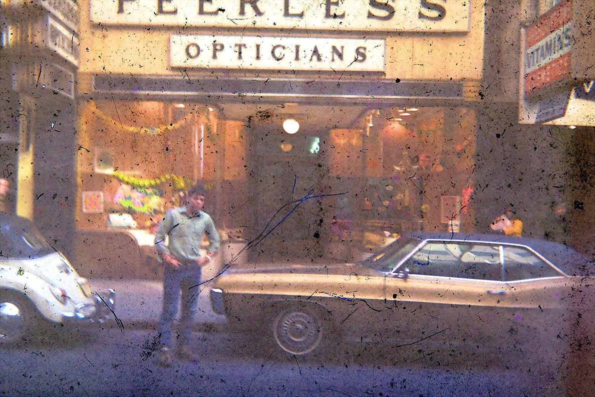 The Peerless Opticians storefront lit up at night, with parked cars and a passer-by on the sidewalk.