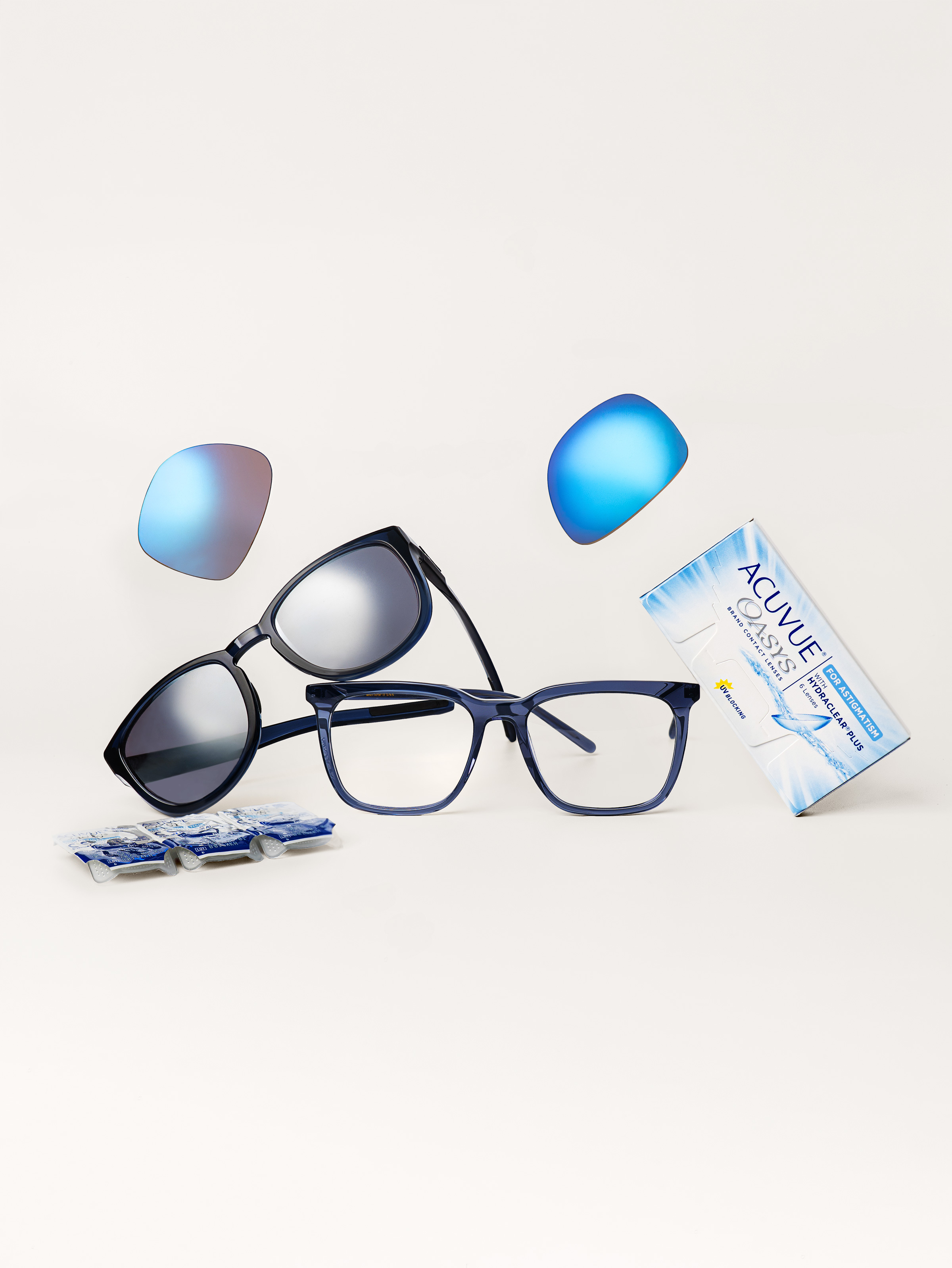 A pair of eyeglasses and stacked sunglasses with mirrored lenses, replacement lens inserts, and a box of Acuvue Oasys contact lenses with a strip of blister packs