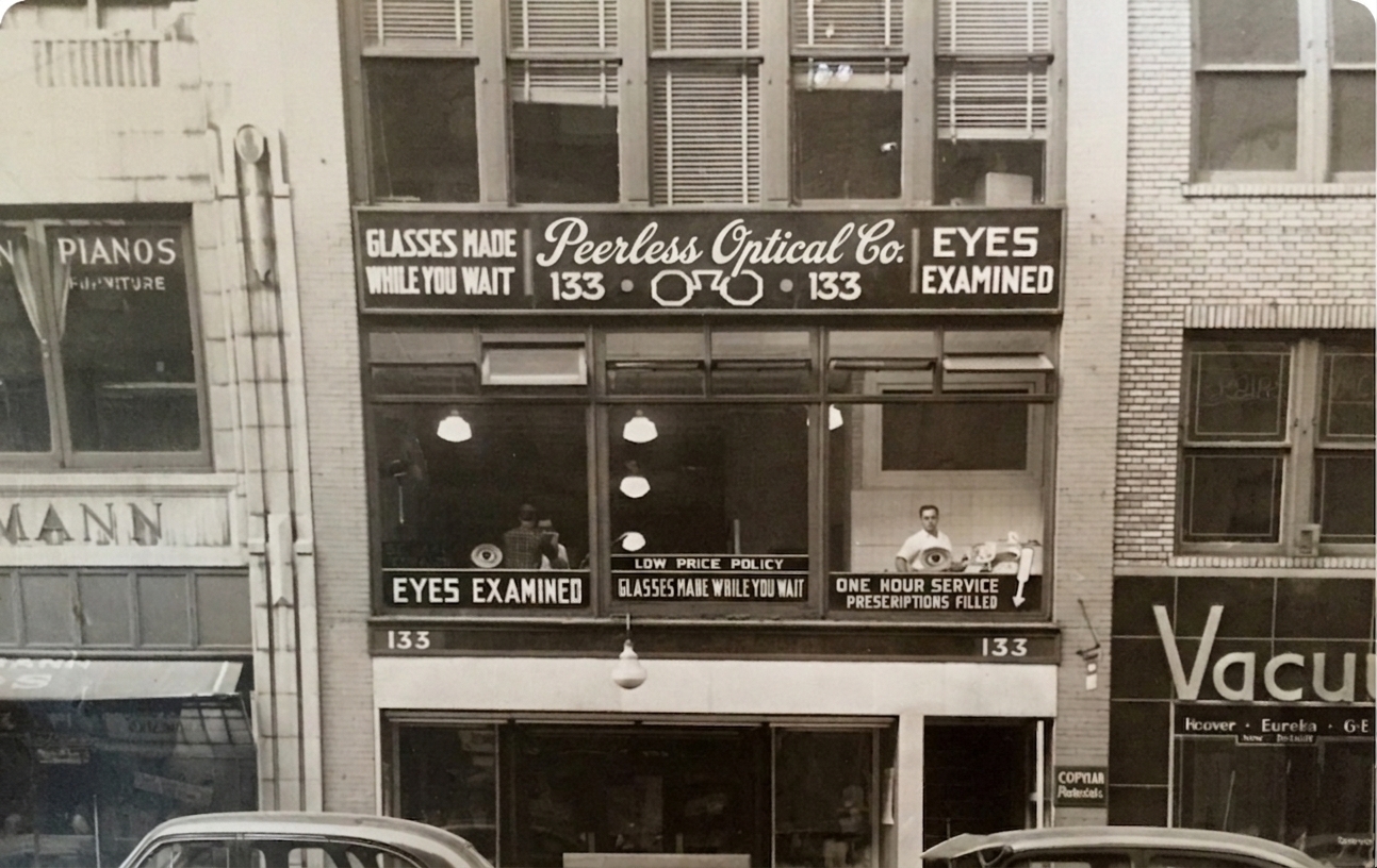 The Peerless Optical Co. storefront, its windows painted with “Glasses made while you wait” and “Eyes examined”.