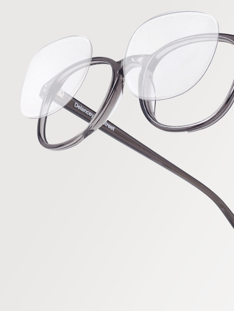 Close-up of translucent gray round eyeglass frames featuring detached, floating lenses positioned above the rims, creating an illusion of suspended optics.